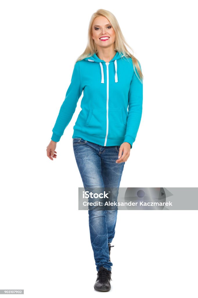 Smiling Casual Blond Woman Is Looking Towards Camera Smiling blond woman in turquoise hoodie and jeans is walking towards camera. Full length studio shot isolated on white. Walking Stock Photo