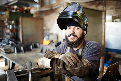 Smiling handsome young craftsman in welding helmet leaning on constriction frame and looking at camera