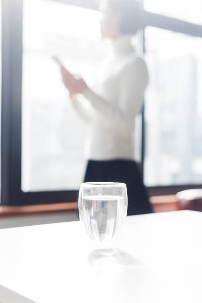 portrait of young woman using phone by window,with a glass of water on foreground,backlit.