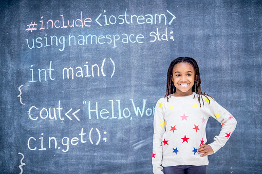 A girl of African descent is taking a programming class in her elementary school. She is standing in front of a blackboard that she has written code on.