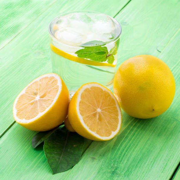 istock Lemonade in a glass, a lemon half, fresh leaves on the green table. A refreshing cold drink of water with ice, mint and slices of lemon, side view 903253124