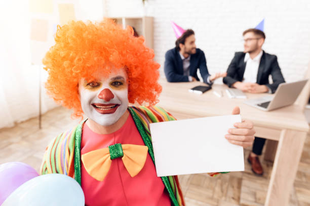 un uomo con un abito da clown e una parrucca è seduto nell'ufficio affari il 1° aprile. - clown laptop bizarre men foto e immagini stock