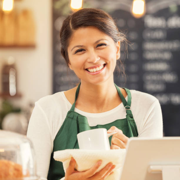 Friendly mid adult female barista in coffee shop Attractive mid adult Asian female barista holds a coffee cup as she works behind the counter in a coffee shop. She is smiling cheerfully at the camera. town criers stock pictures, royalty-free photos & images