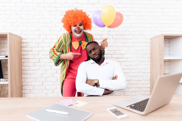 un uomo con un abito da clown è in piedi accanto a un uomo di colore che si siede alla sua scrivania. - clown laptop bizarre men foto e immagini stock