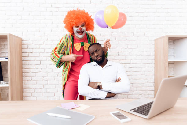 道化師のスーツの男は、彼の机に座っている黒人男性の横に立っています。 - clown laptop bizarre men ストックフォトと画像