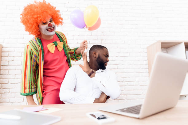 un uomo con un abito da clown è in piedi accanto a un uomo di colore che si siede alla sua scrivania. - clown laptop bizarre men foto e immagini stock