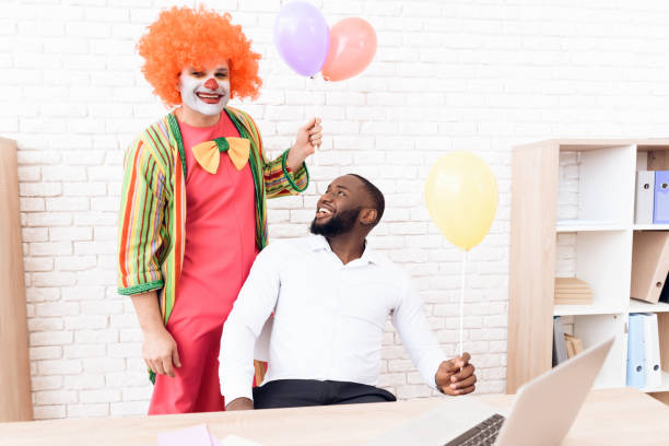 un uomo con un abito da clown è in piedi accanto a un uomo di colore che si siede alla sua scrivania. - clown laptop bizarre men foto e immagini stock