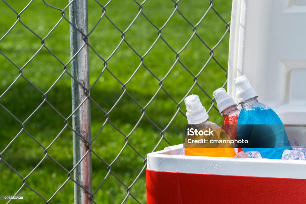 Ice cold colorful sports drinks on ice in cooler on a hot day Various colors and flavors of ice cold sports drinks in a red and white ice chest with a chainlink fence behind it on a hot Summer day with shadow from a tree on the grass in the background. This could be a public park or sports field. Sport Drink Stock Photo