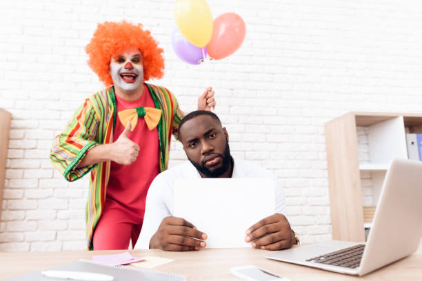 un uomo con un abito da clown è in piedi accanto a un uomo di colore che si siede alla sua scrivania. - clown laptop bizarre men foto e immagini stock