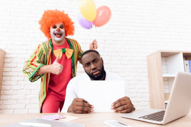 道化師のスーツの男は、彼の机に座っている黒人男性の横に立っています。 - clown laptop bizarre men ストックフォトと画像