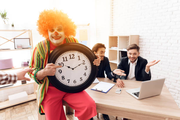 un uomo con un abito da clown sta accanto agli uomini in abiti da lavoro, che siedono alla scrivania. - clown laptop bizarre men foto e immagini stock