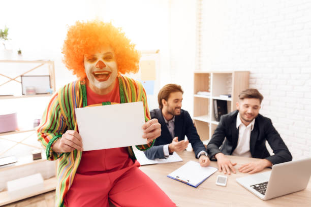 un uomo con un abito da clown sta accanto agli uomini in abiti da lavoro, che siedono alla scrivania. - clown laptop bizarre men foto e immagini stock