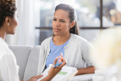 Mid adult counseling patient has a confused expression on her face while listening to a counselor. The counselor is gesturing while talking with the patient.