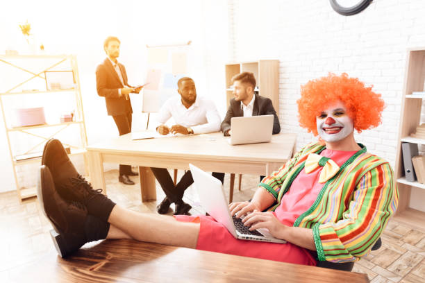 un uomo con un vestito da clown seduto alla sua scrivania, i piedi sul tavolo. - clown laptop bizarre men foto e immagini stock