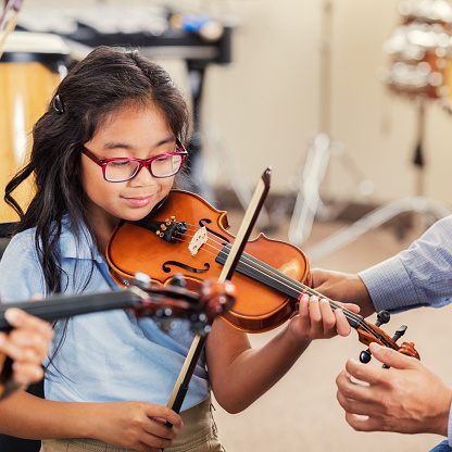 A smiling elementary age girl looks down as she plays the violin in music class at school.  An unrecognizable teacher reaches toward her to help with proper positioning.