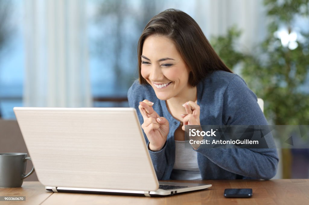 Happy girl crossing fingers checking on line content Happy hopeful girl crossing fingers checking on line content in a laptop at home Internet Stock Photo