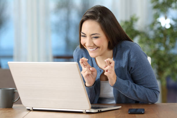 Happy girl crossing fingers checking on line content Happy hopeful girl crossing fingers checking on line content in a laptop at home auction photos stock pictures, royalty-free photos & images