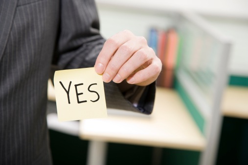 A man in an office setting wearing a suit is holding up a yellow Post-It note that says, 