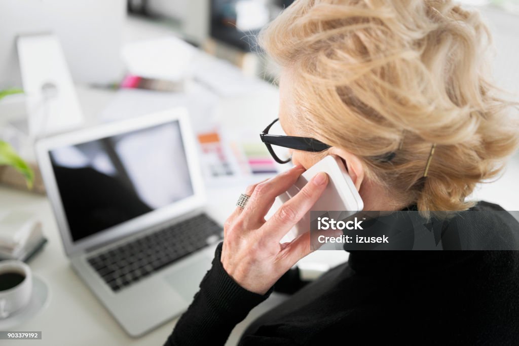 Senior businesswoman working on laptop in the office Senior businesswoman sitting at the desk in the office, using laptop and talking on phone. High angle view. 60-69 Years Stock Photo