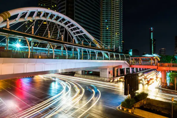 Beautiful and colorful light trails of the vehicles under the skywalk at night at the intersection on Sathorn road in bustling Bangkok, Thailand with city's skyscrapers in the background