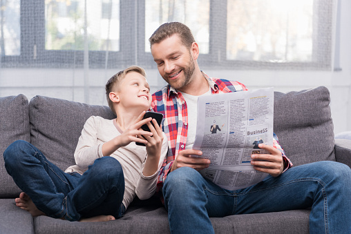smiling father with newspaper and cute little son using smartphone together at home
