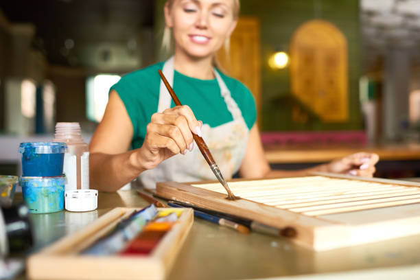 Portrait of pretty blonde woman enjoying work in art studio painting shutters with bronze paint, focus on foreground