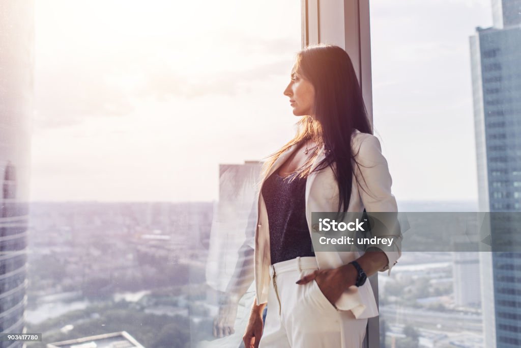 Retrato de dama elegante negocio usar traje formal blanco de pie junto a la ventana mirando el paisaje urbano - Foto de stock de Mujeres libre de derechos