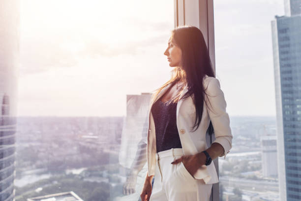 retrato de dama elegante negocio usar traje formal blanco de pie junto a la ventana mirando el paisaje urbano - home interior young adult professional occupation vertical fotografías e imágenes de stock