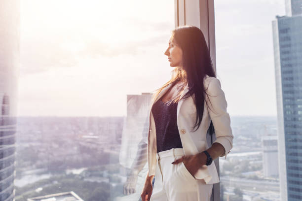 retrato de mulher de negócios elegante, vestindo terno branco formal em pé perto da janela, olhando a paisagem urbana - executive apartment - fotografias e filmes do acervo