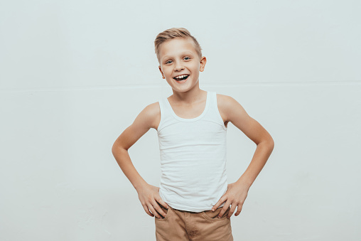 Young smiling boy in white tank top standing with hands on hips and looking at camera, isolated on white