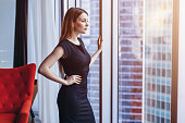 Well-off attractive woman thinking standing at the window admiring cityscape in her penthouse apartment