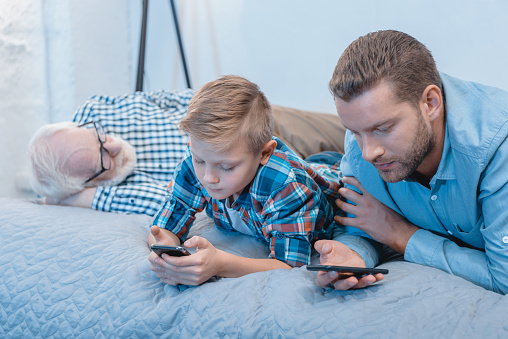 Little boy and his father lying on bed using smartphones while grandpa is sleeping