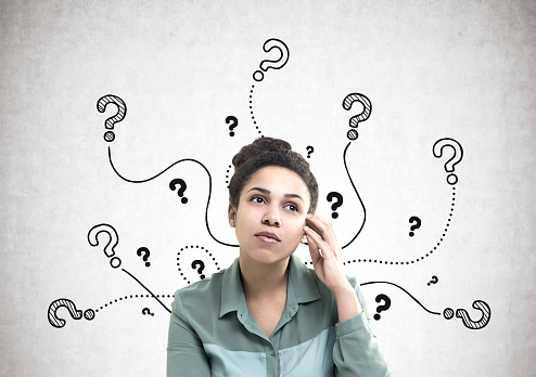 Portrait of a young and pensive African American woman wearing a green shirt and looking upwards. She is standing near a concrete wall with many question marks on it.