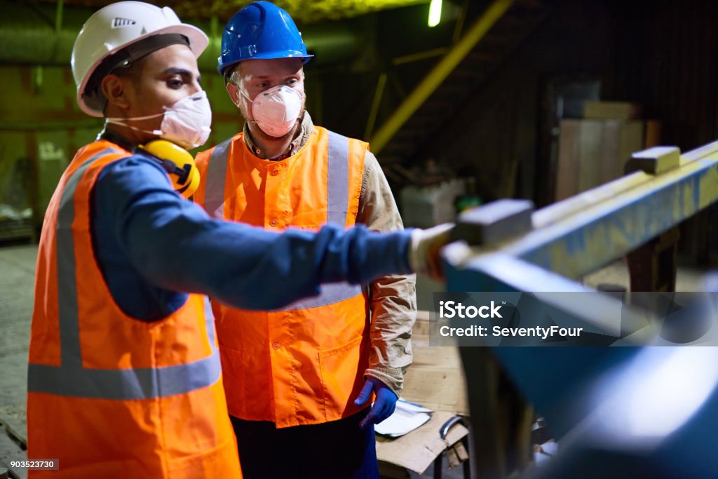 Discussing Results of Joint Work with Colleague Handsome mixed race technician wearing reflective vest pointing at something while discussing results of joint work with his colleague, interior of spacious production department on background Manufacturing Occupation Stock Photo