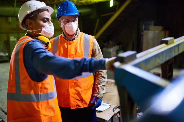 Discussing Results of Joint Work with Colleague Handsome mixed race technician wearing reflective vest pointing at something while discussing results of joint work with his colleague, interior of spacious production department on background protective mask workwear stock pictures, royalty-free photos & images
