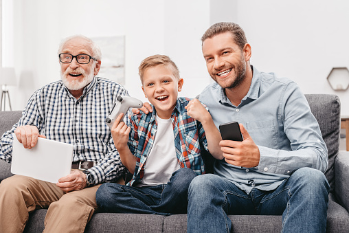 Father, son and grandfather sitting together on couch in living room holding digital tablet, smartphone and gamepad