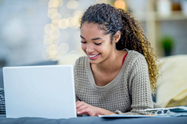 Girl Using A Laptop A teenage girl of African descent is indoors in her bedroom. She is wearing casual clothes. She is laying in bed with her laptop, and smiling while typing on the keyboard. 13 14 years child teenage girls lying down stock pictures, royalty-free photos & images