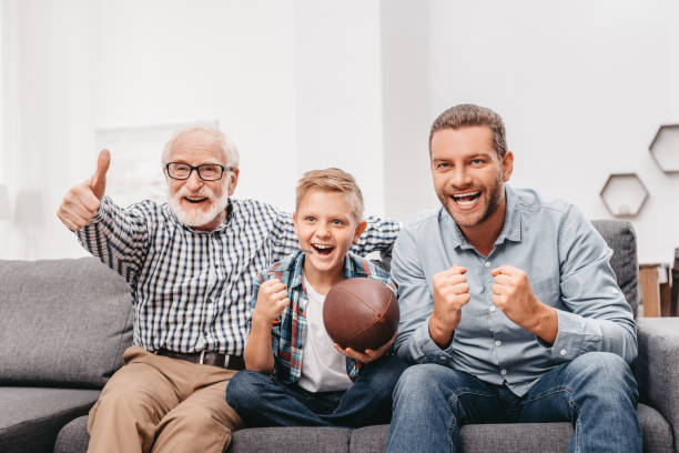 Family watching football at home Little boy on couch with grandfather and father, cheering for a football game and holding a football ball old football stock pictures, royalty-free photos & images