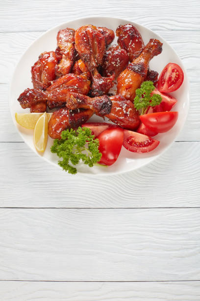 tasty grilled crispy juicy teriyaki chicken tasty grilled crispy juicy teriyaki chicken drumsticks sprinkled with sesame seeds on white plate with tomatoes, parsley and lemon slices, vertical view from above sticky sesame chicken sauces stock pictures, royalty-free photos & images