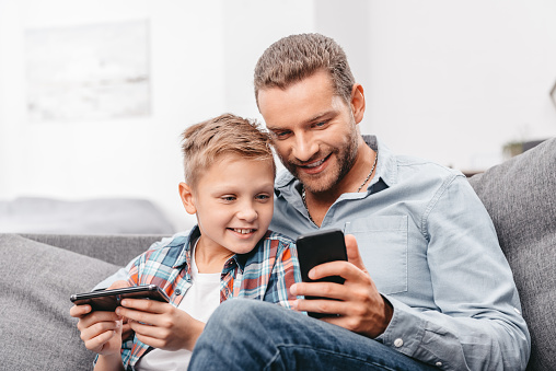 Father and little son sitting on couch in living room and using smartphones