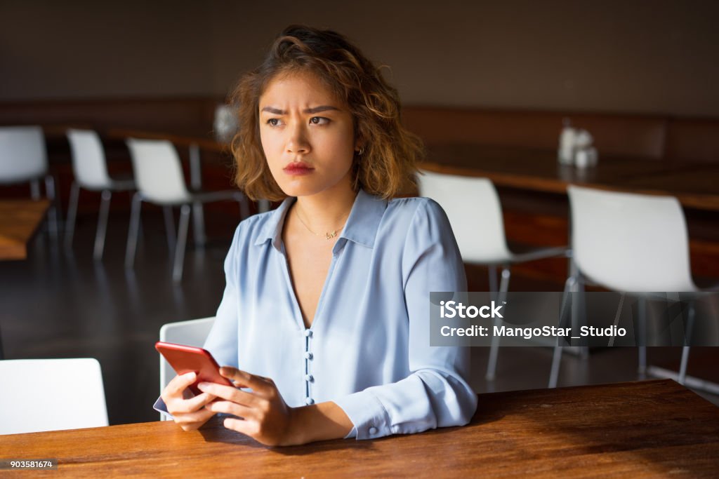 Severe Young Businesswoman with Phone at Cafe Closeup of strict young Asian woman sitting at table in cafe and holding mobile phone. Frowning business woman chatting and getting bad news. Communication and bad news concept Adult Stock Photo