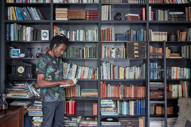 Mature man reading book by shelves Mature man reading book by shelves. Male is standing at home. He is wearing casuals. book display stock pictures, royalty-free photos & images
