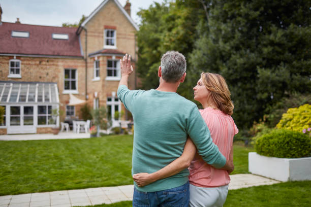 Mature couple standing in front of house Rear view of mature couple standing in front of house. Man and woman are with arms around in lawn. They are planning with architecture in background. english spoken stock pictures, royalty-free photos & images