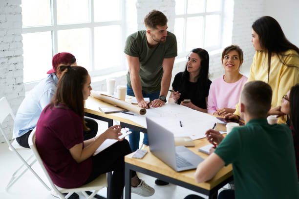 istock Handsome bearded male coach checking accountings and graphic planning for architectural project during training workshop in college with dream team of young students in modern loft interior office 903624056