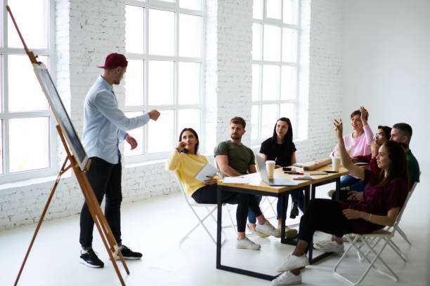 istock Young creative casually dressed male hipster moderating discussion of startup projects during conference with skilled marketing experts, teacher asking questions on lesson with group of students 903624104