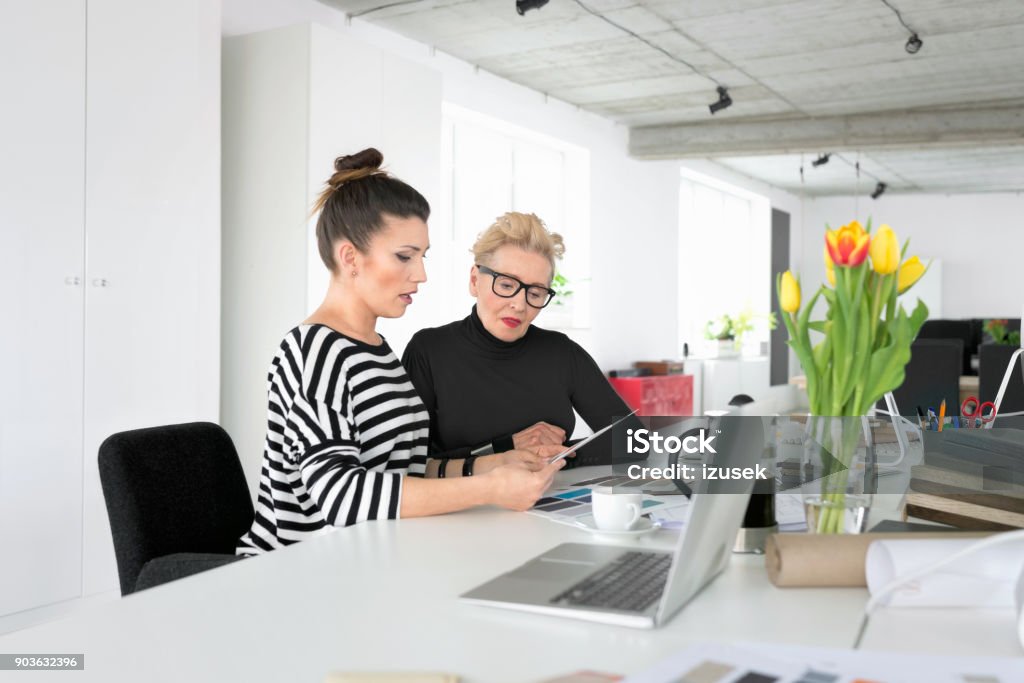 Senior and young interior designers using a digital tablet in the office Senior interior designer working together with young woman in the office, sitting at the desk and using a digital tablet together. 60-69 Years Stock Photo