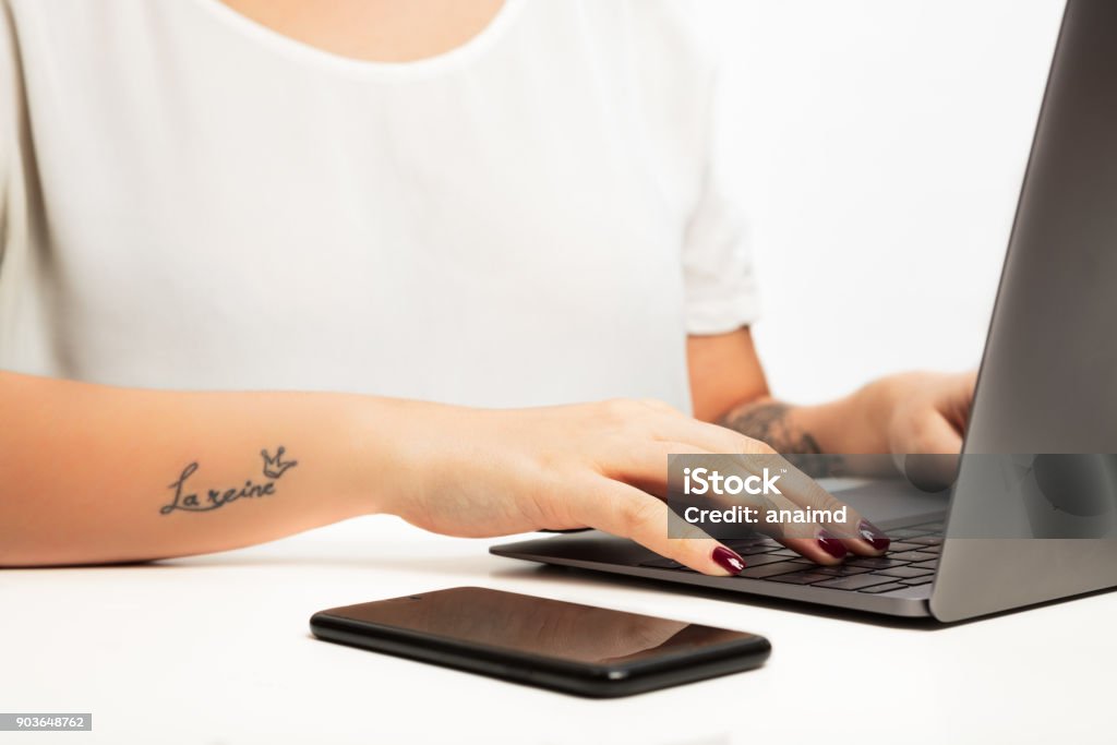 Hands of a young woman typing on a laptop Hands of a young woman typing on a laptop computer in a close up view with a mobile phone alongside. With âla reineâ tattoo on arm, meaning â queenâ  in french  with crown sign Adult Stock Photo