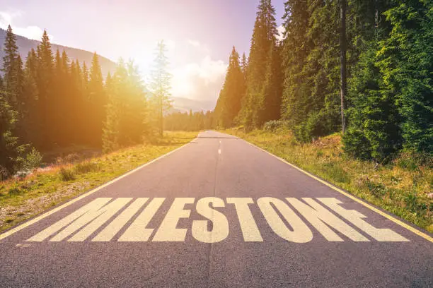 Photo of Asphalt road with arrow guideline and Milestone letters painted on the surface. An image of a road milestones are representative of success in the future goal. Road to success with light of the sun