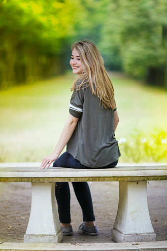 Portrait of a cheeky teenage girl sat on a stone bench laughing and looking over her shoulder with a blurred green background