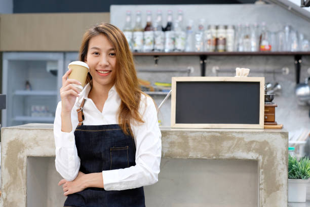 young asia woman barista holding a disposable coffee cup with smiling face at cafe counter background, small business owner, food and drink industry concept - restaurant food food and drink industry food service occupation imagens e fotografias de stock